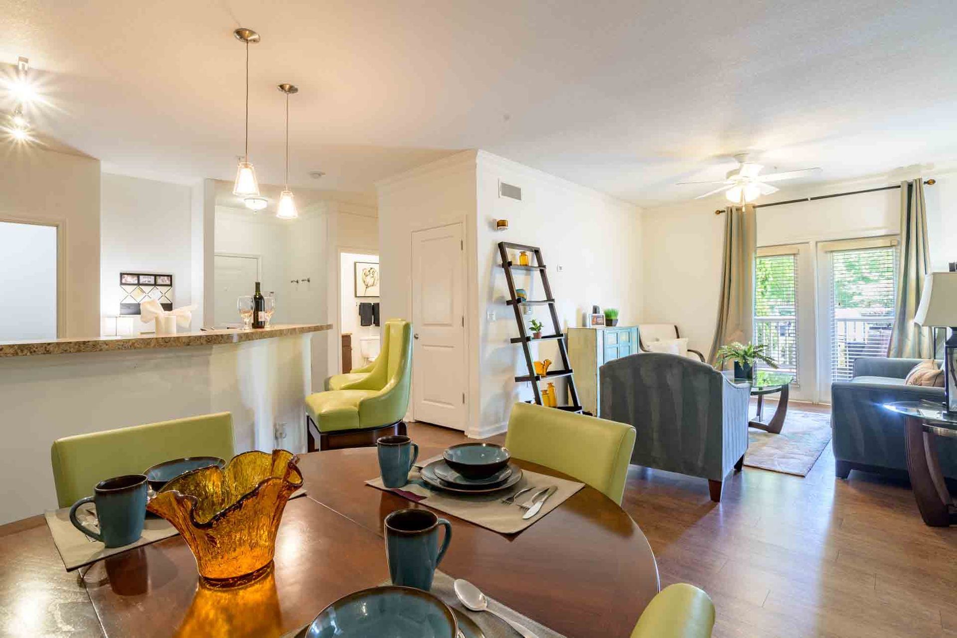 Dining area with round table, adjacent to a living room. Green and blue accents, neutral walls.