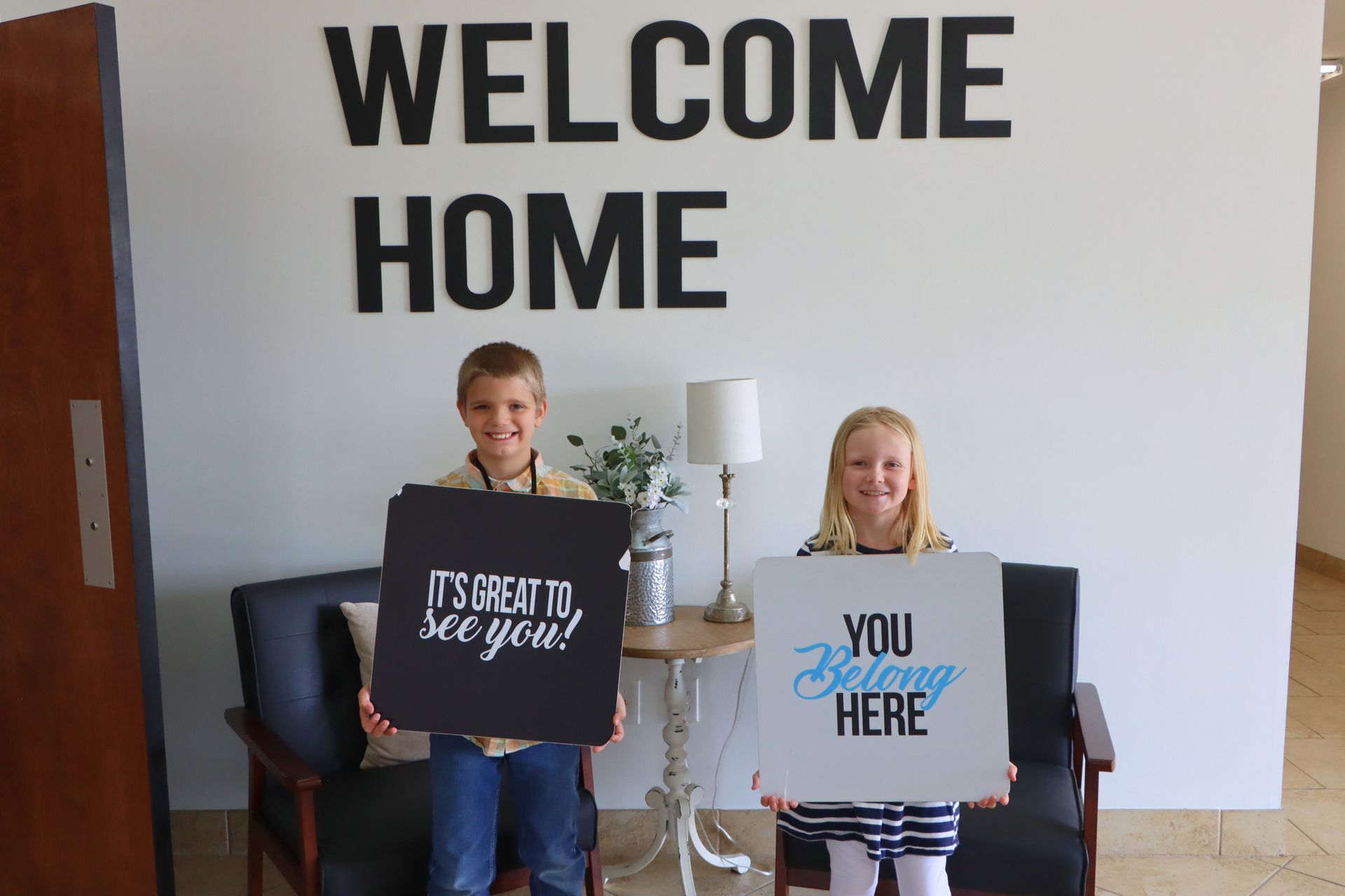 A boy and a girl are holding signs in front of a wall that says welcome home.