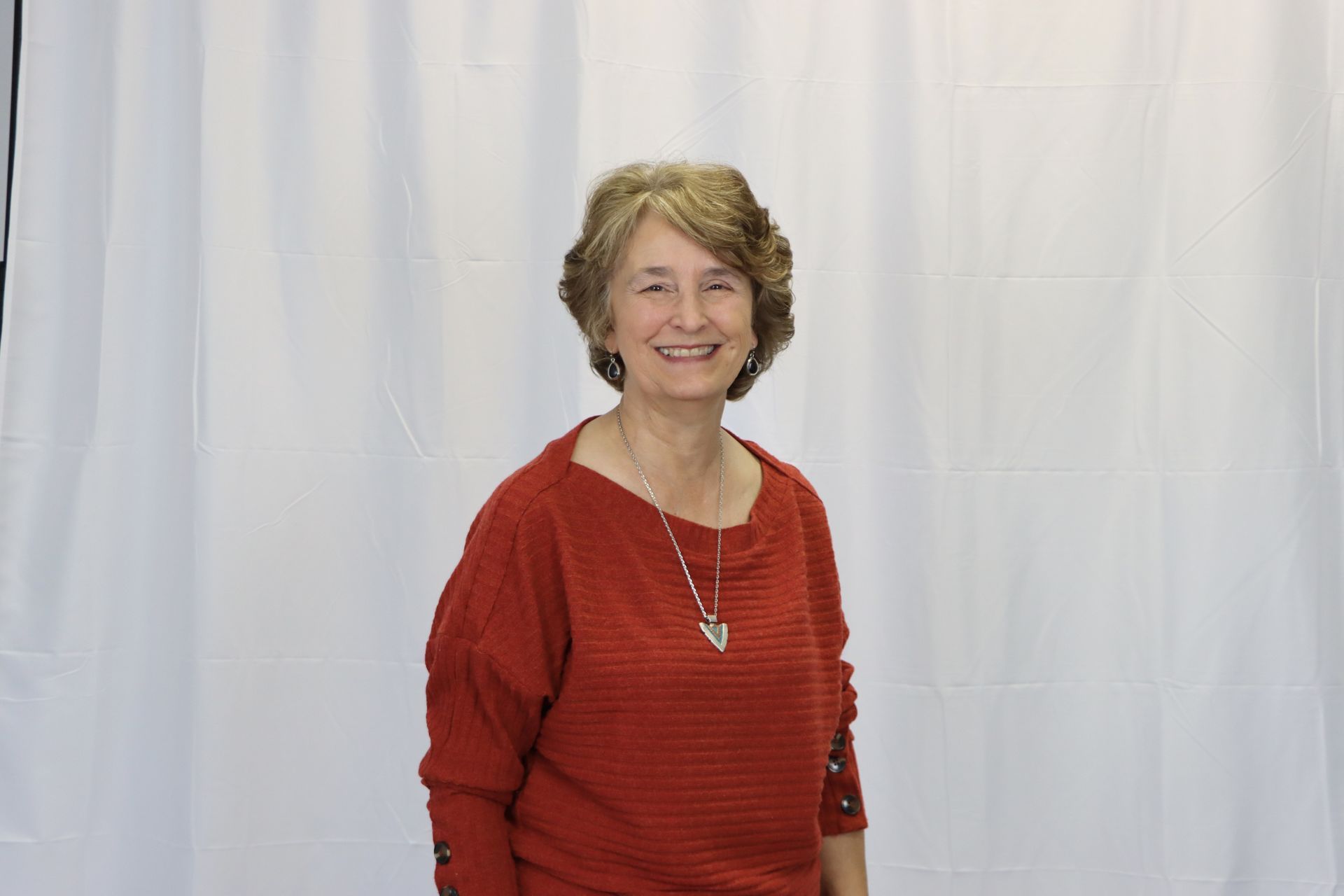 A woman in a red sweater and necklace is smiling in front of a white curtain.