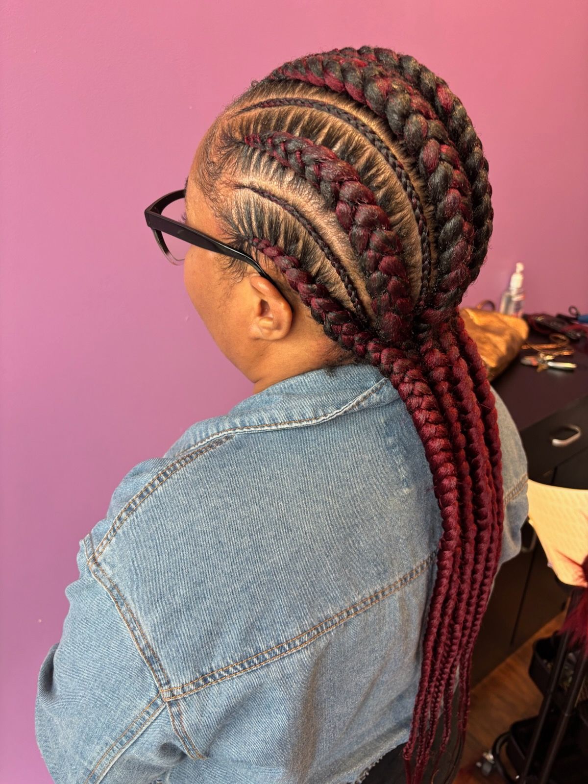 Woman with burgundy and black braided hairstyle, wearing glasses and a denim jacket.