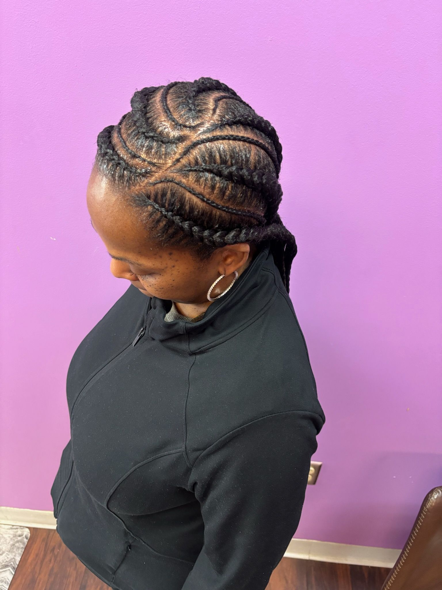 Woman with dark skin and braided hairstyle, wearing a black jacket, standing in front of a purple wall.