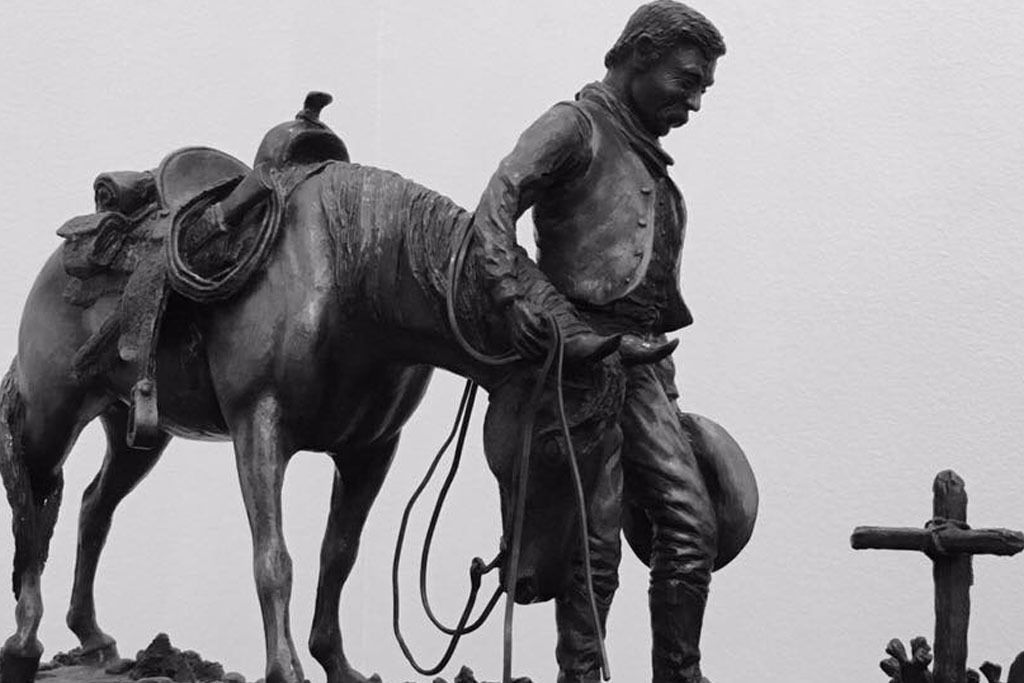 Bronze cowboy statue with saddled horse, rope, and cross in the background.