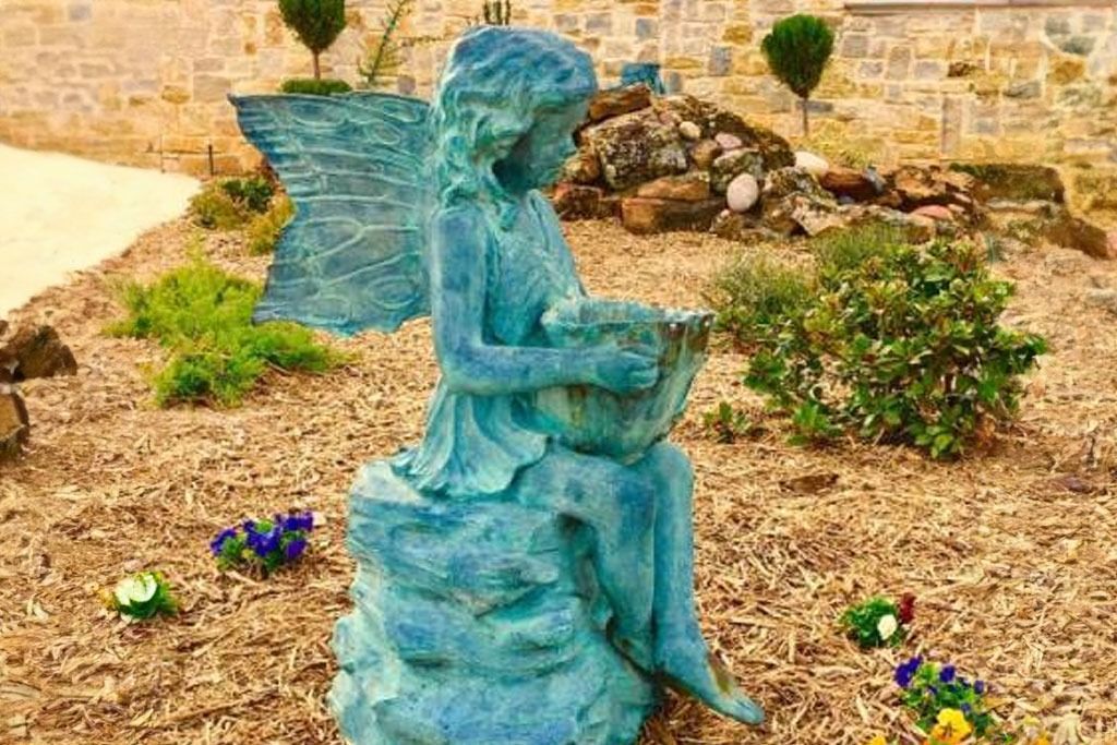 Blue fairy statue sitting in a garden, holding a basket, surrounded by plants and flowers.