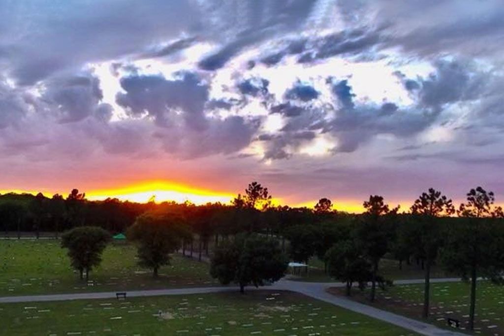 Sunset over a cemetery with green grass, trees, and cloudy sky.