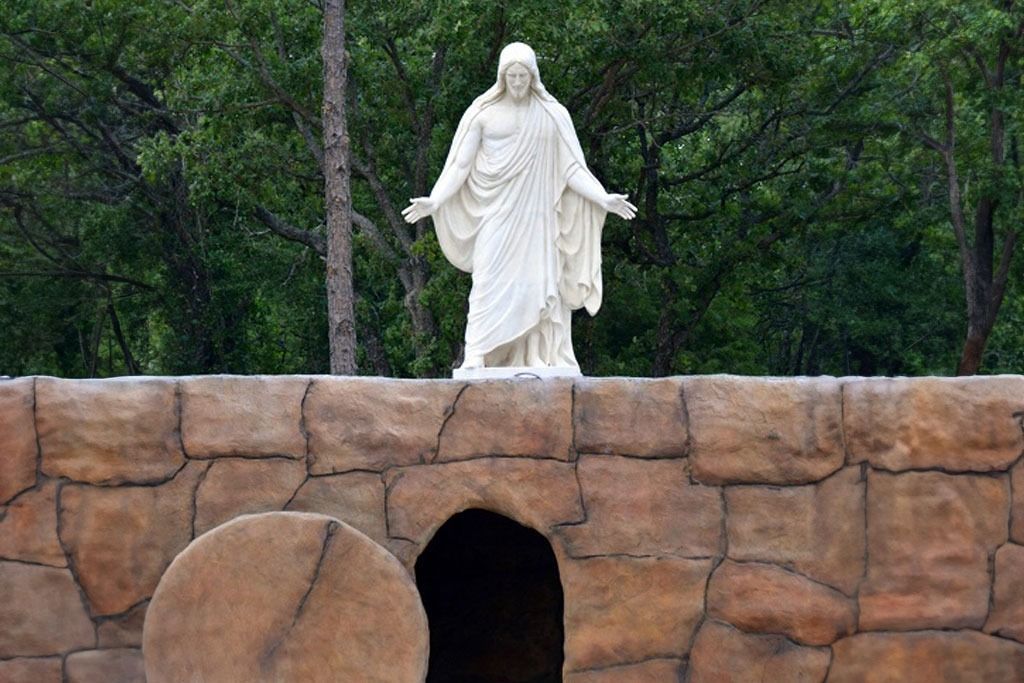 White statue of Jesus with open arms above a tomb and brown stone wall, with green trees in the background.
