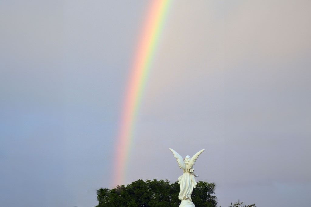 Rainbow arcs over a white angel statue with outstretched wings, set against a cloudy sky.