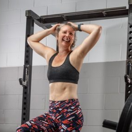 Woman in workout clothes smiles, posing near weight rack in a gym.