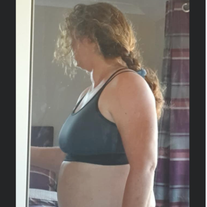 Woman in black sports bra stands by a mirror. She has curly hair in a braid, facing to the side.