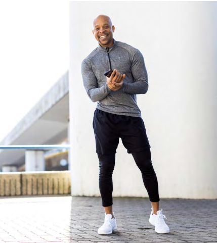 Man in athletic wear smiles, holding phone outdoors, by a white pillar.