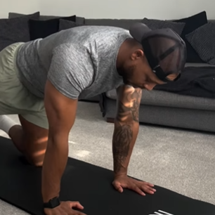Man on hands and knees on a black mat, wearing a gray shirt and cap, exercising indoors.