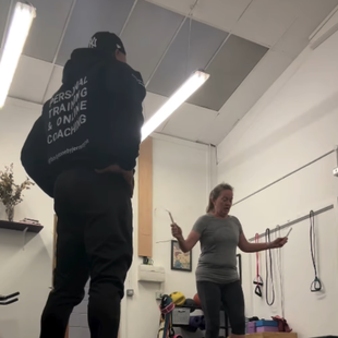 A person in a black sweatshirt watches a woman jump rope in a gym setting.