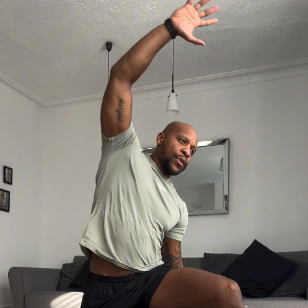 Man in gray shirt and black shorts stretching arm upwards in a living room.