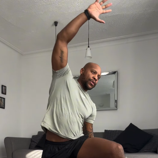 Man in gray shirt and black shorts stretching arm upwards in a living room.