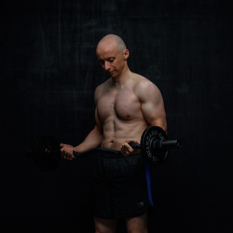 Muscular man curls a barbell, indoors. He's shirtless, wearing shorts. Black background.