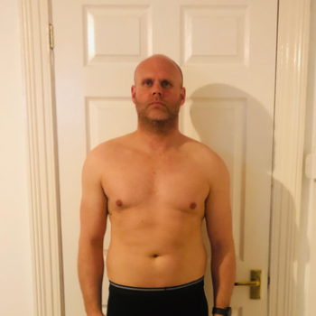 Man shirtless, standing in front of a white door. He has short hair and a neutral expression.