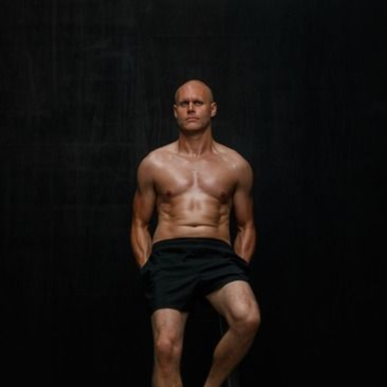 Shirtless man in black shorts, standing with hands behind his back, against a dark background.
