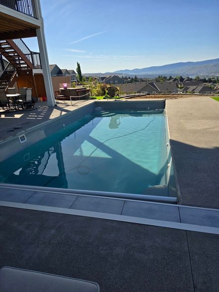 A rectangular swimming pool with blue water, grey concrete surround. Houses and mountains in the distance.