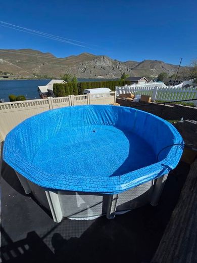 Above-ground pool covered with a blue tarp, overlooking a lake and mountains on a sunny day.