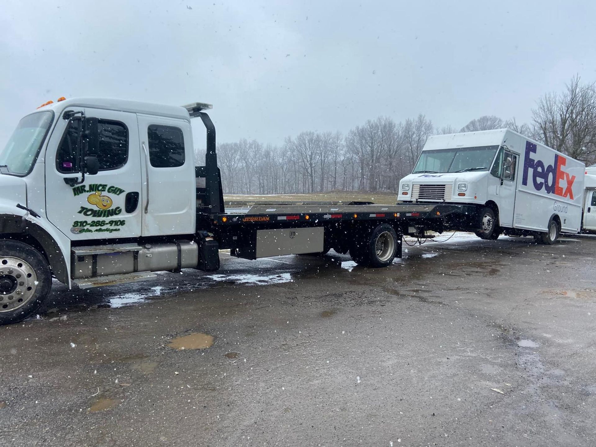 A tow truck is towing a fedex truck in a parking lot.