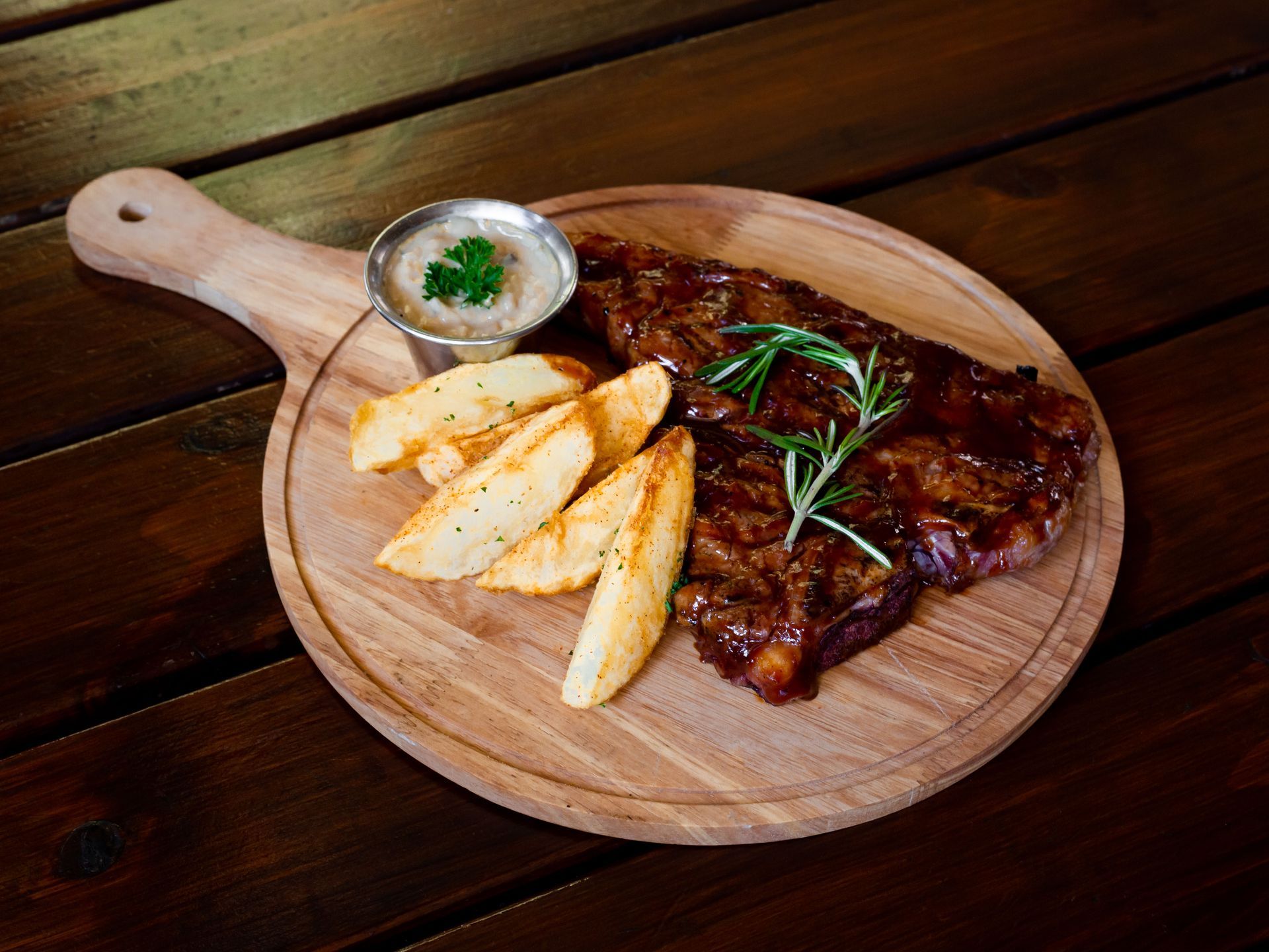 A steak and potato wedges on a wooden cutting board on a wooden table.
