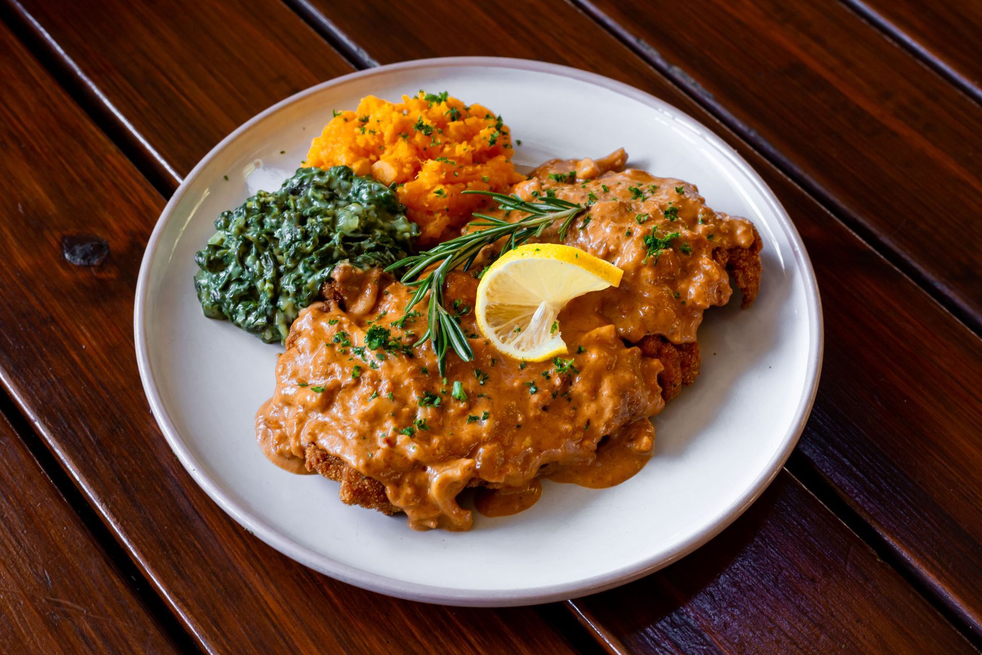 A white plate topped with meat , mashed potatoes and spinach on a wooden table.