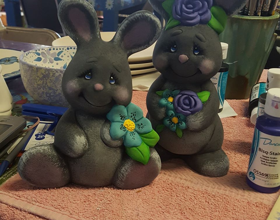Two bunny figurines sit on a table next to a bottle of purple paint