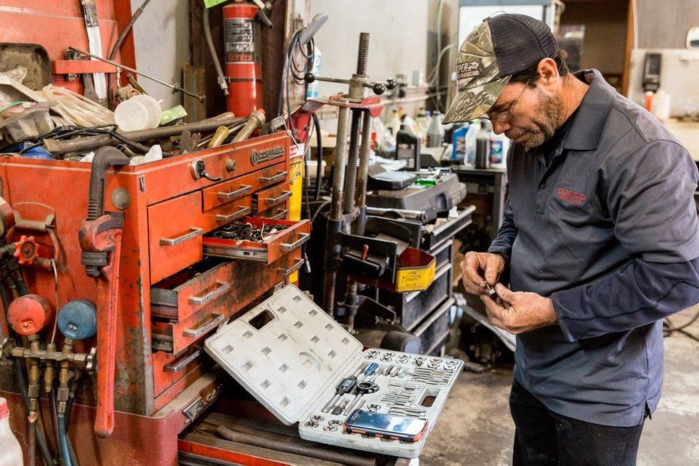 Mechanic in garage with tools, examining parts, wearing a camouflage cap.