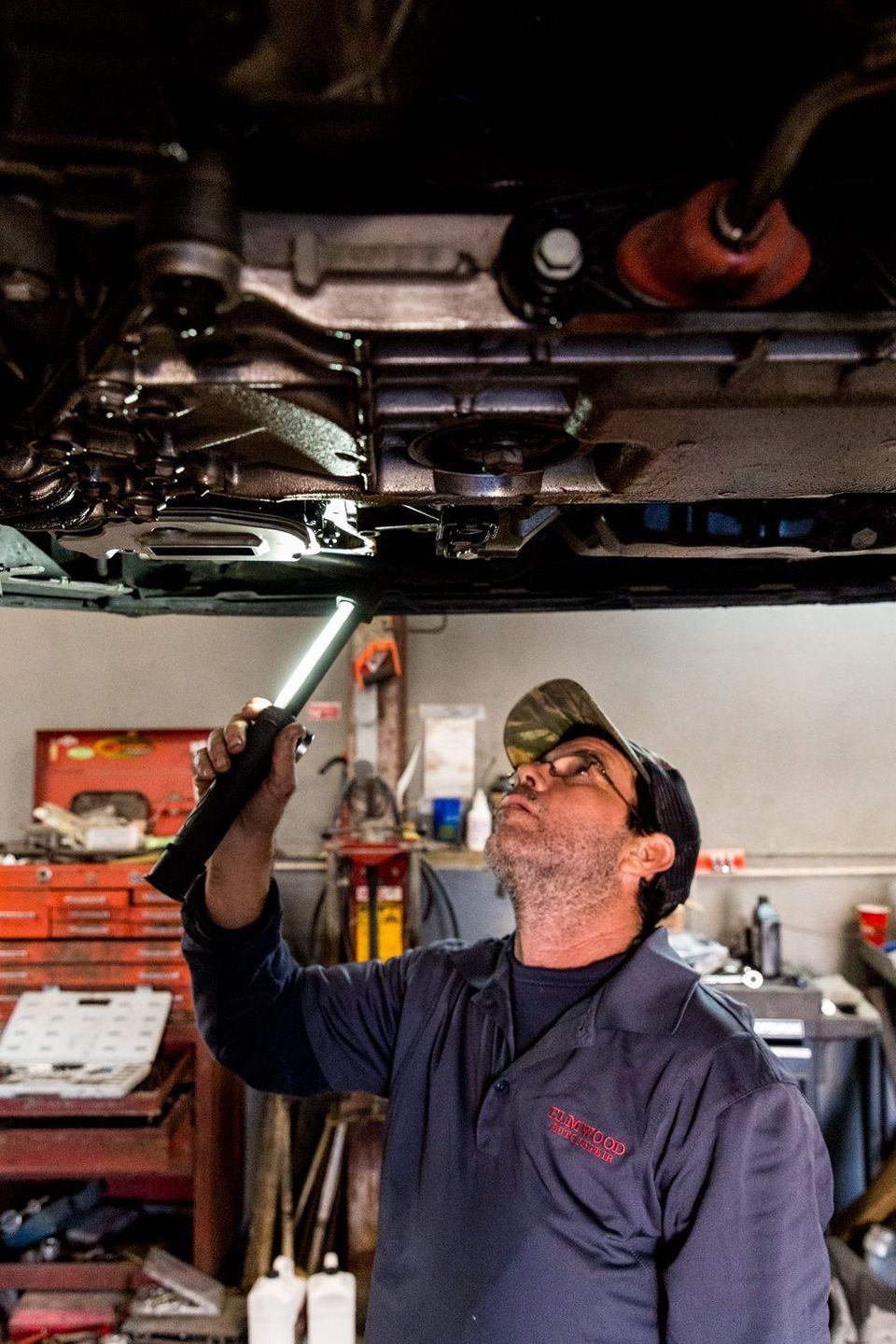 Mechanic inspecting car underside with flashlight. Wearing blue coveralls, looking up. Workshop setting.