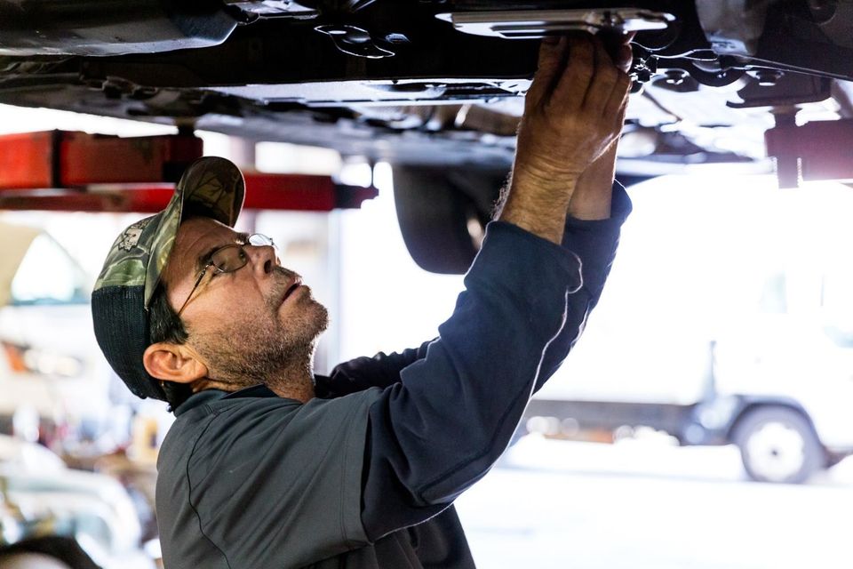 Mechanic under a car, wearing a cap and glasses, working on the vehicle in a garage.