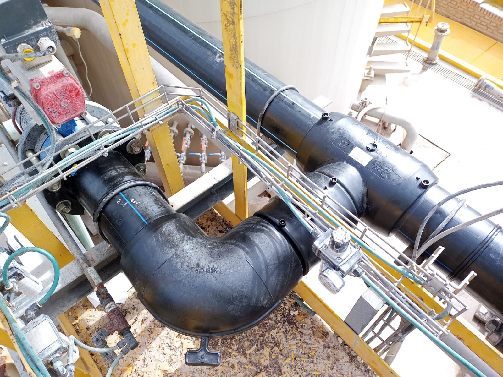 Tubería industrial de color negro con conexión en codo sobre estructura metálica de color amarillo.