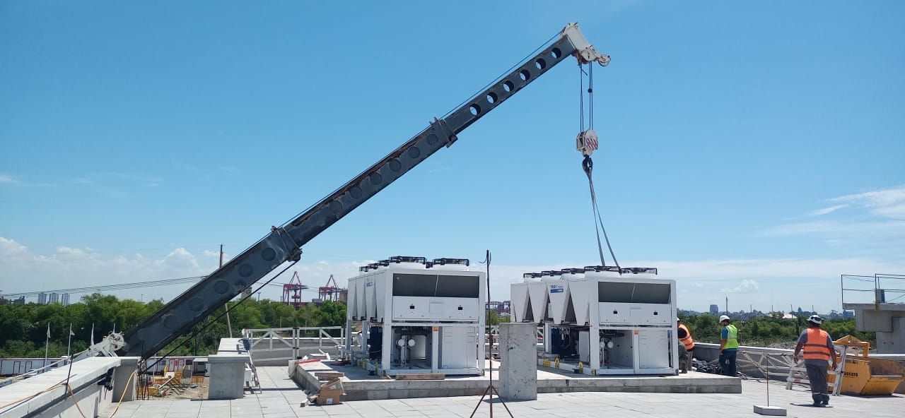 Una grúa levanta dos grandes unidades industriales blancas sobre un tejado bajo un cielo azul claro.