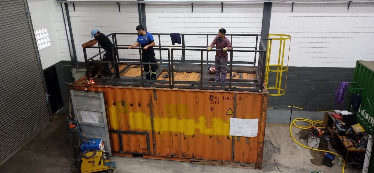 Trabajadores soldando en un contenedor de envío naranja con una plataforma en un taller.