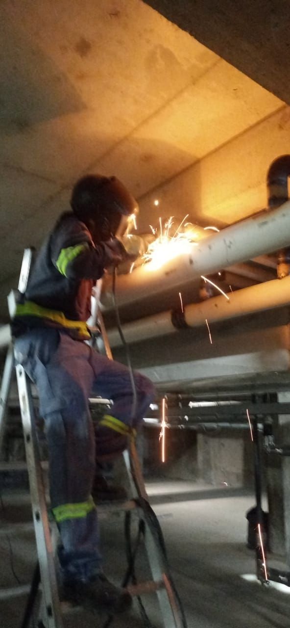 Soldador con equipo de seguridad en una escalera, trabajando con tuberías en un entorno de hormigón. Saltan chispas.