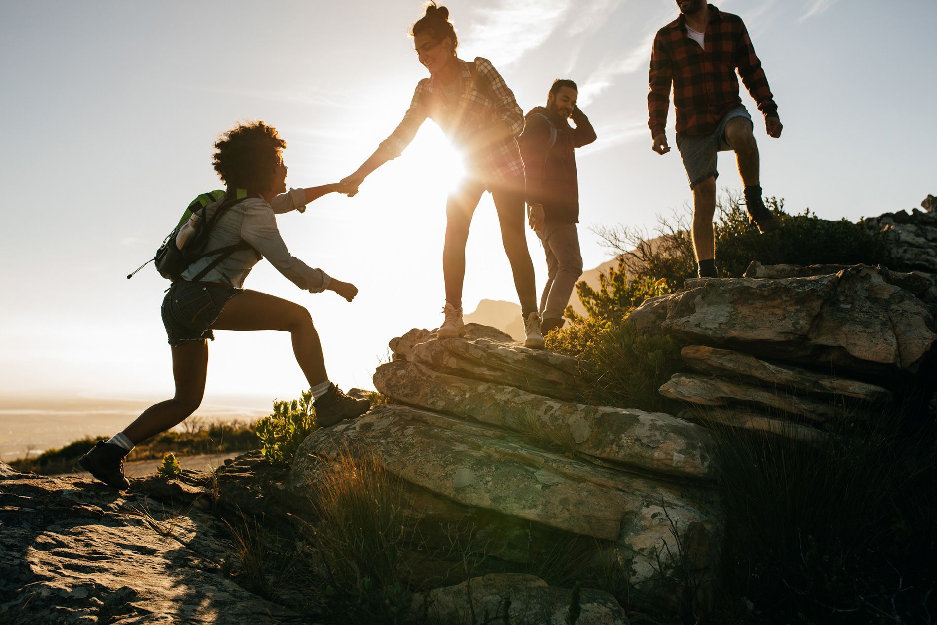Hikers helping each other climb rocky terrain, sunlit scene.