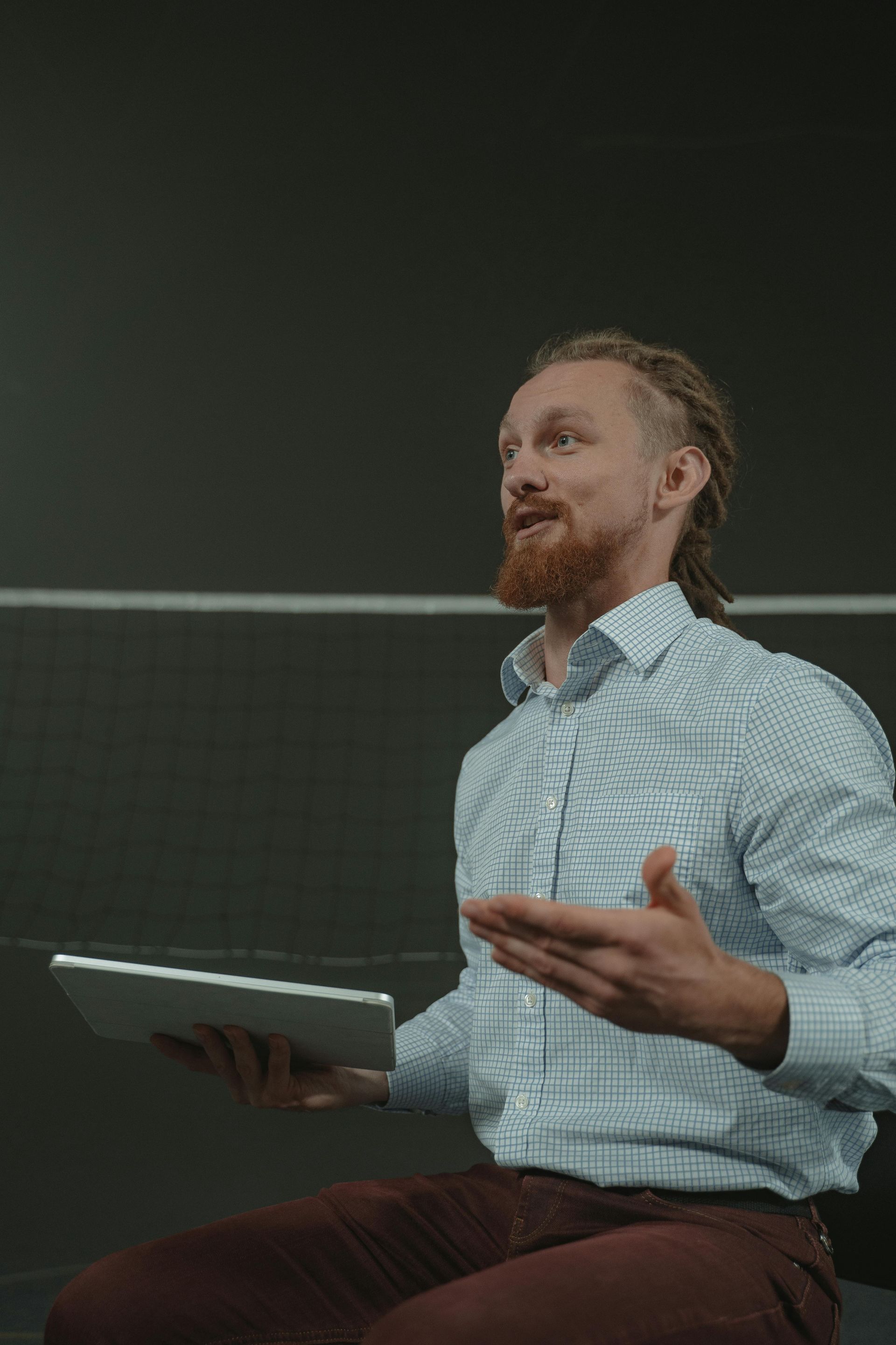 Man with dreadlocks gesturing while holding tablet, seated in front of a chalkboard.