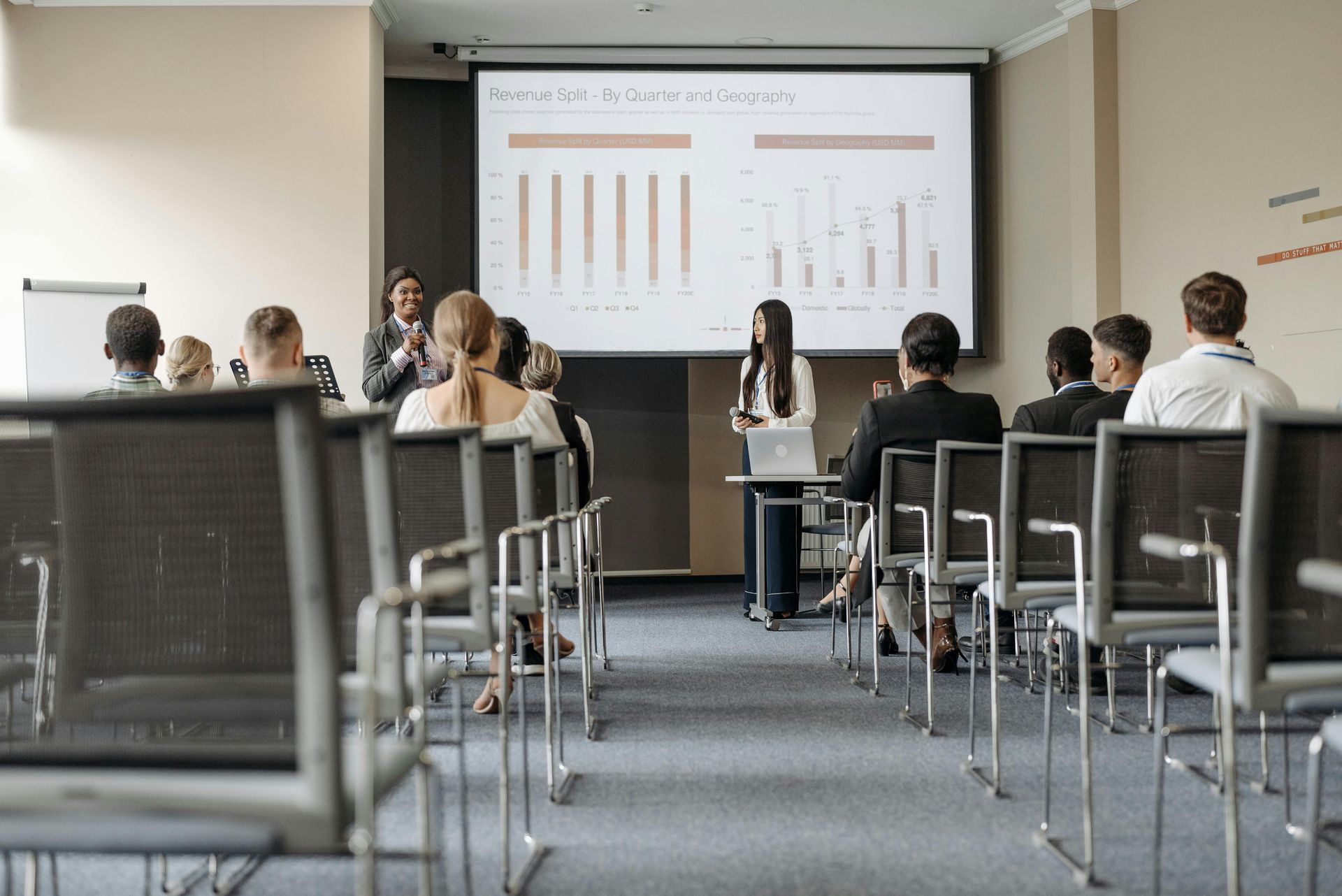 A woman presents data on a screen in a conference room to an audience seated in rows of chairs.