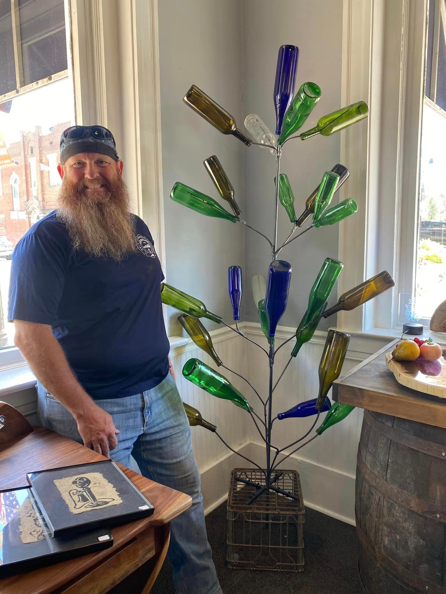 A man with a beard is standing in front of a christmas tree made of wine bottles.