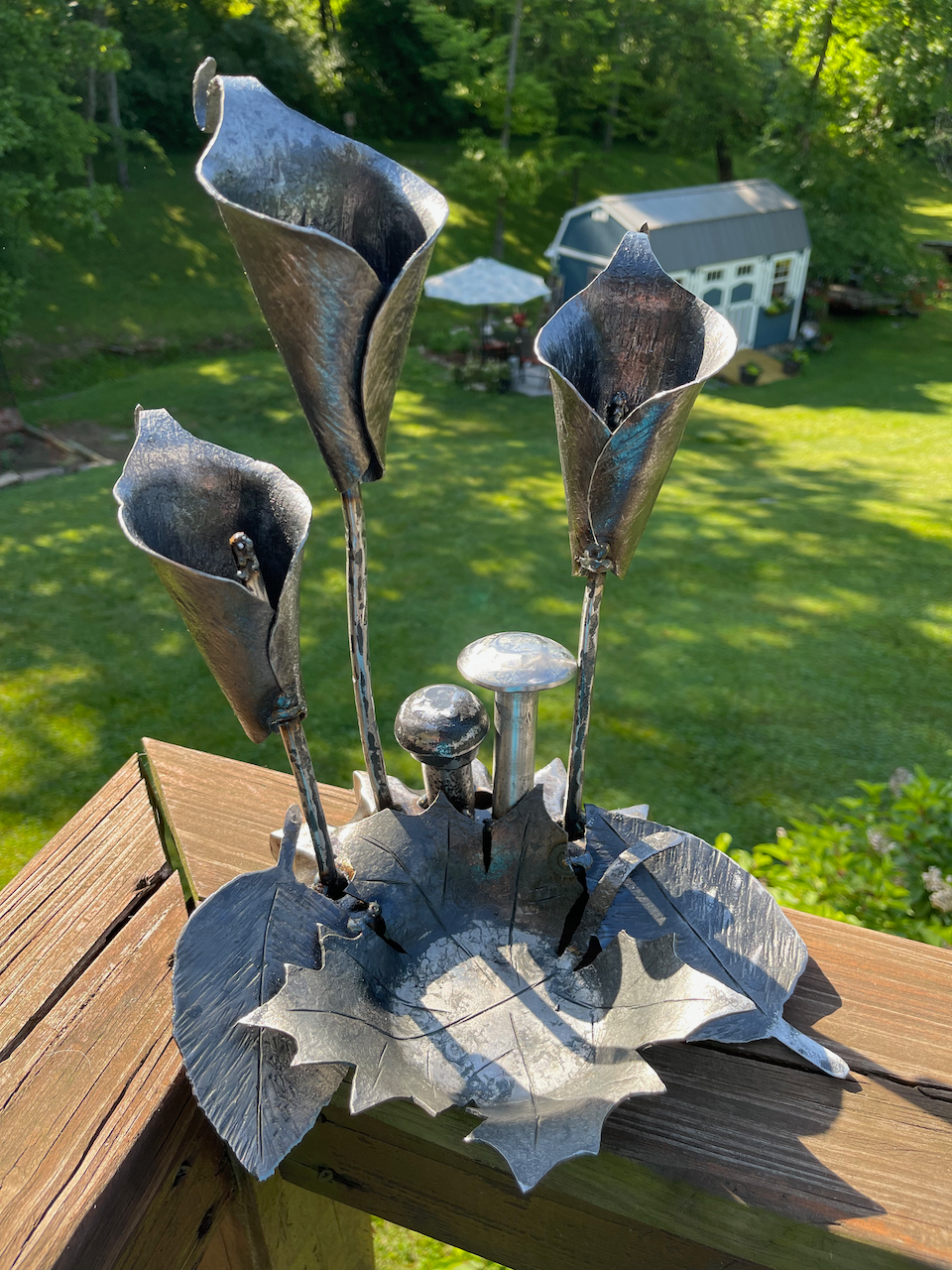 A metal sculpture of flowers and mushrooms is sitting on a wooden deck.
