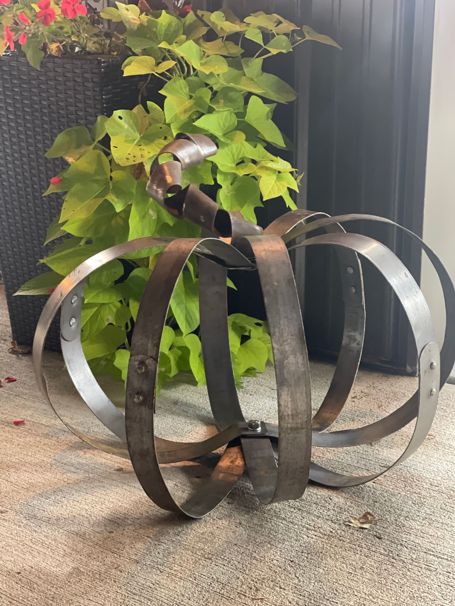 A metal pumpkin is sitting on the ground in front of a potted plant.