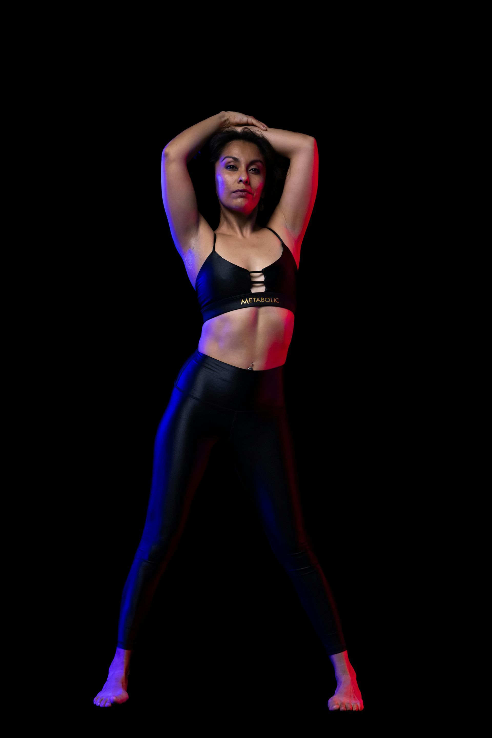 Mujer vestida con ropa deportiva negra con los brazos levantados, iluminada por luz azul y roja, sobre un fondo negro.