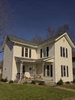 Two-story white house with black shutters, front porch, and green lawn against a blue sky.
