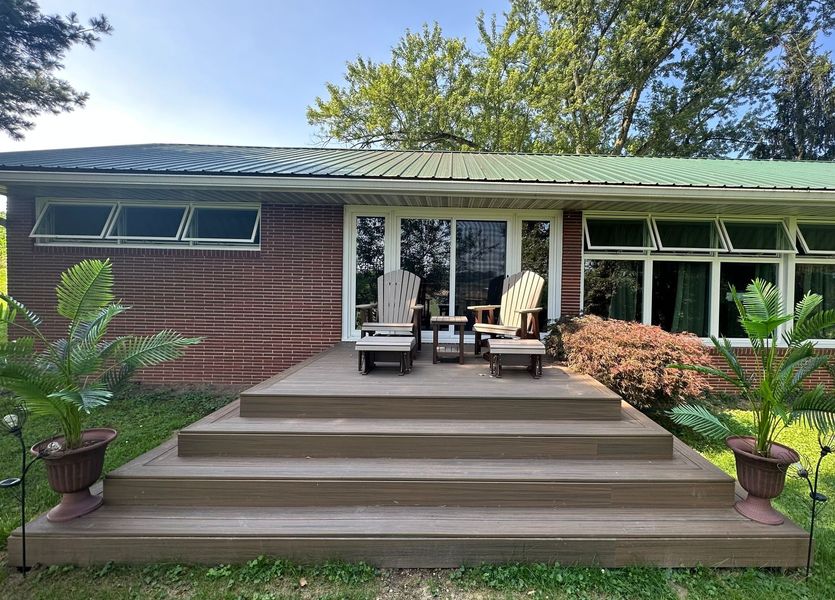 A brown wooden deck with chairs in front of a house with a green roof and large windows, flanked by potted plants.