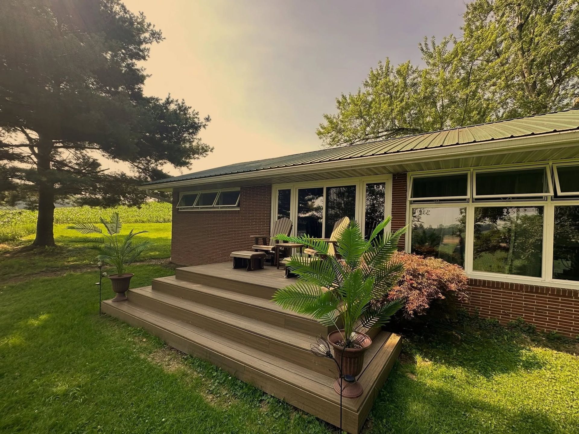 A brick house with a wooden deck, large windows, and a surrounding green lawn and trees.