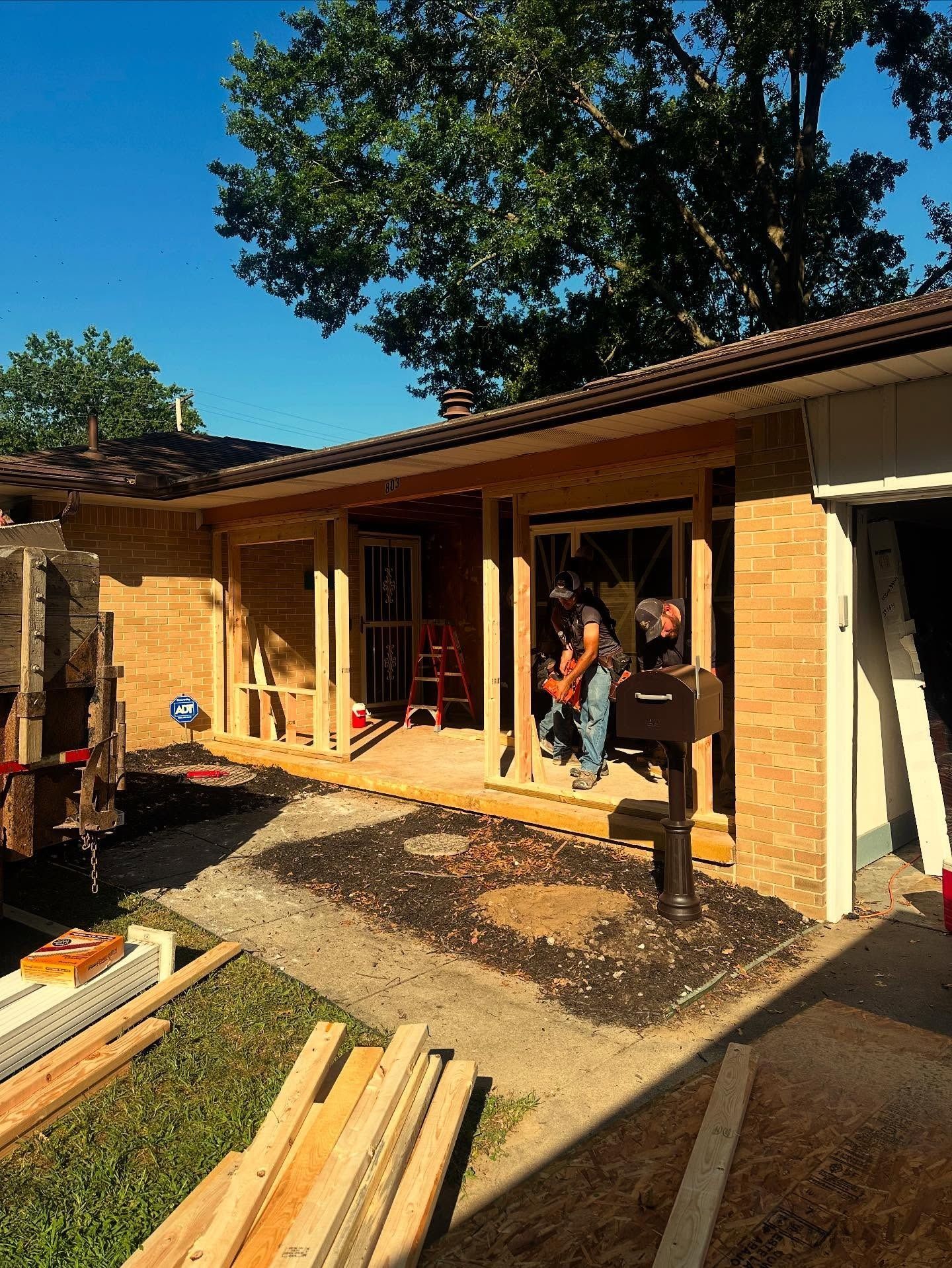 Construction site: porch addition. A person saws wood. Brown wood, blue sky, green lawn, and gravel.