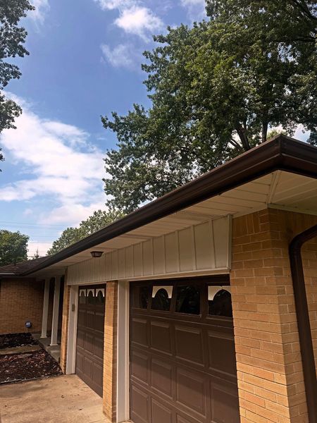 Brown garage doors beneath a brown roofline. Beige paneling, brick wall, and a partly cloudy sky are visible.