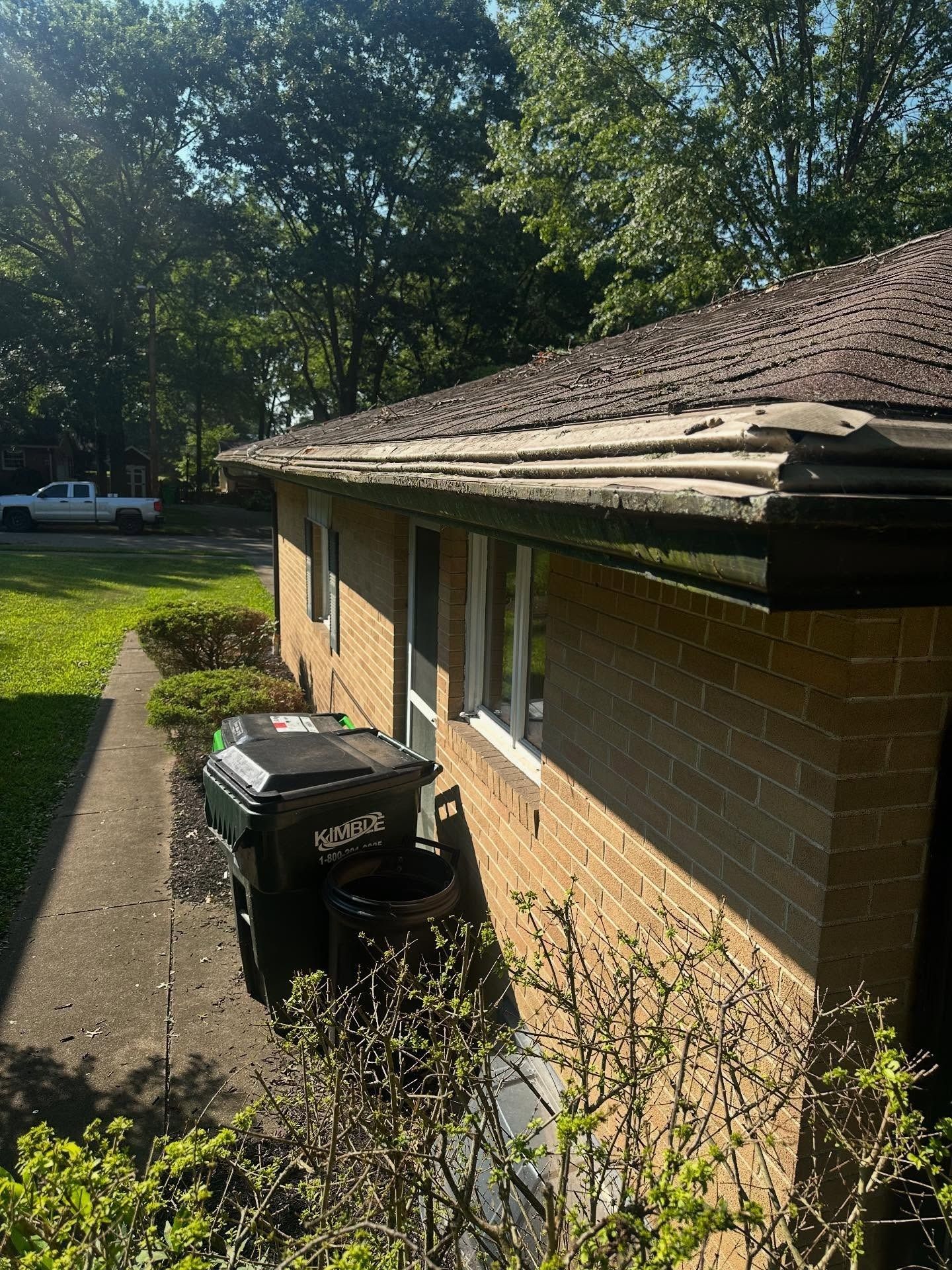 A brick house with a damaged roof and overgrown gutters. A trash bin is visible.