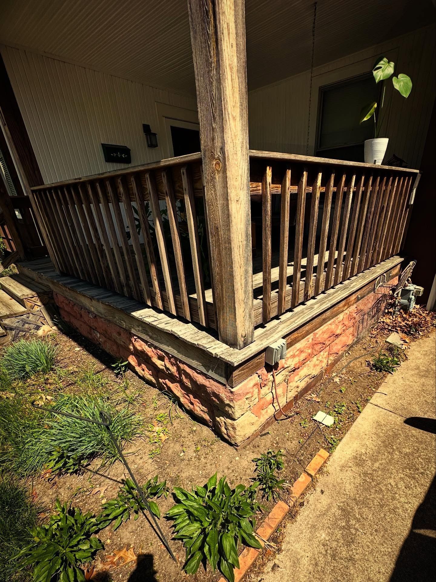 Wooden porch with brick base, surrounded by dirt and plants.