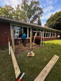 Two men building a fence in front of a brick house with large windows on a sunny day.