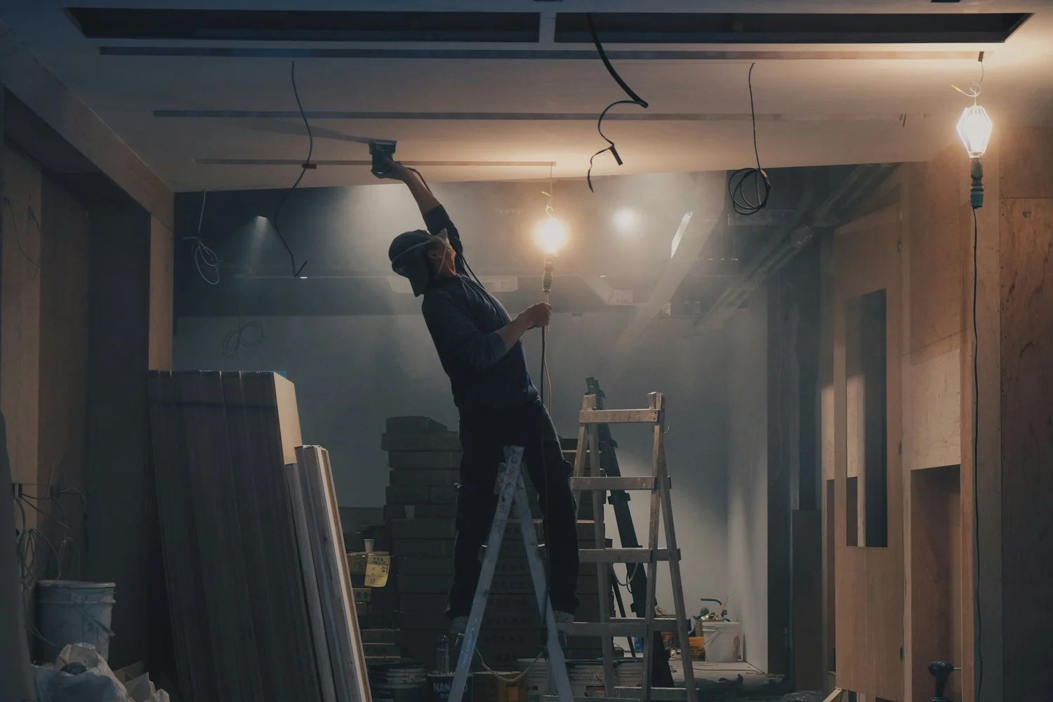 Construction worker on a ladder, reaching up to work on ceiling electrical wires in a dusty room.