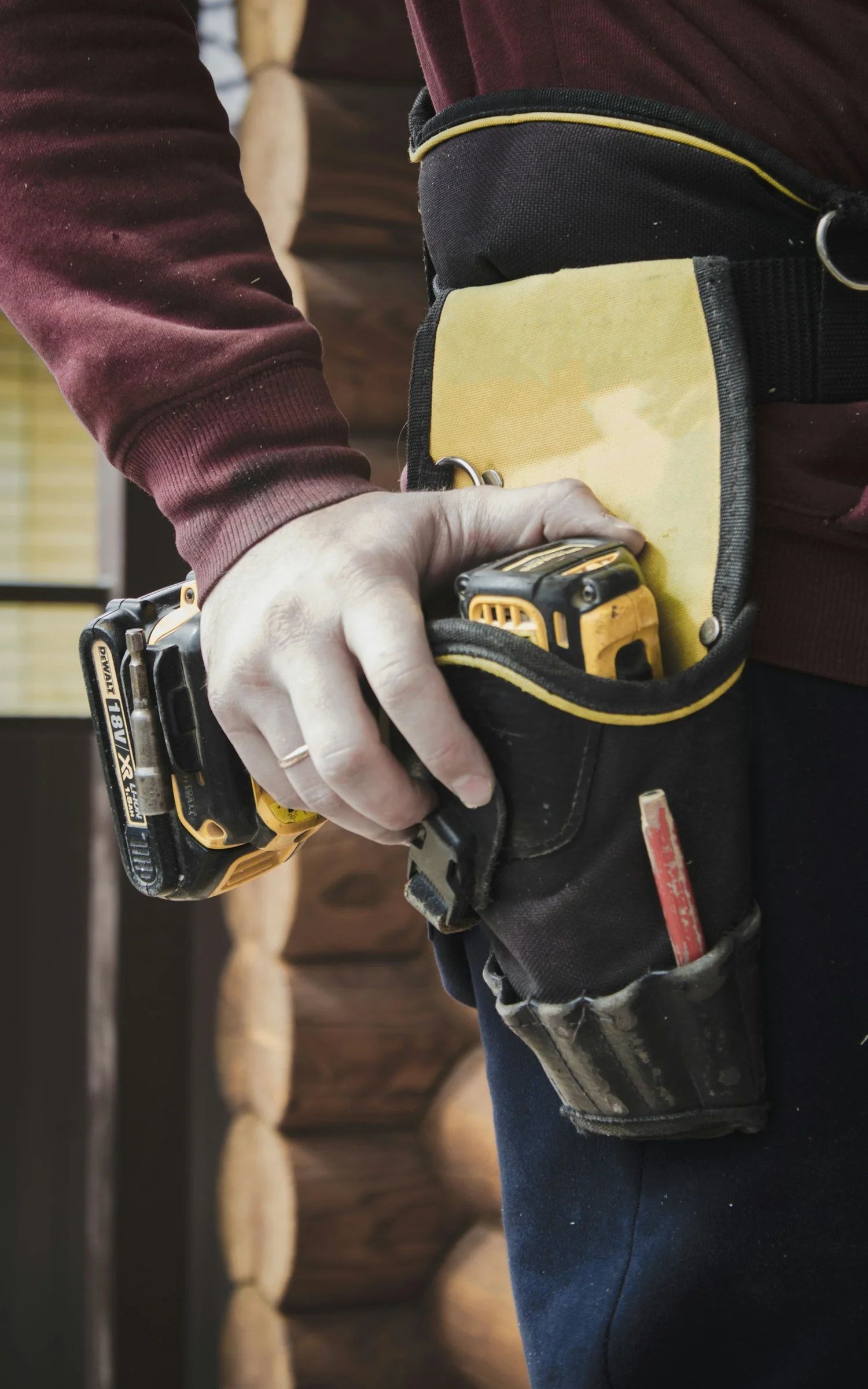 Person wearing a black tool belt with a drill and holding a tape measure. Red pencil visible.